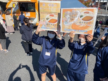 ２年４組のお店：勝華麗（カツカレー）のカレーショップの看板を手に呼び込みする生徒さん